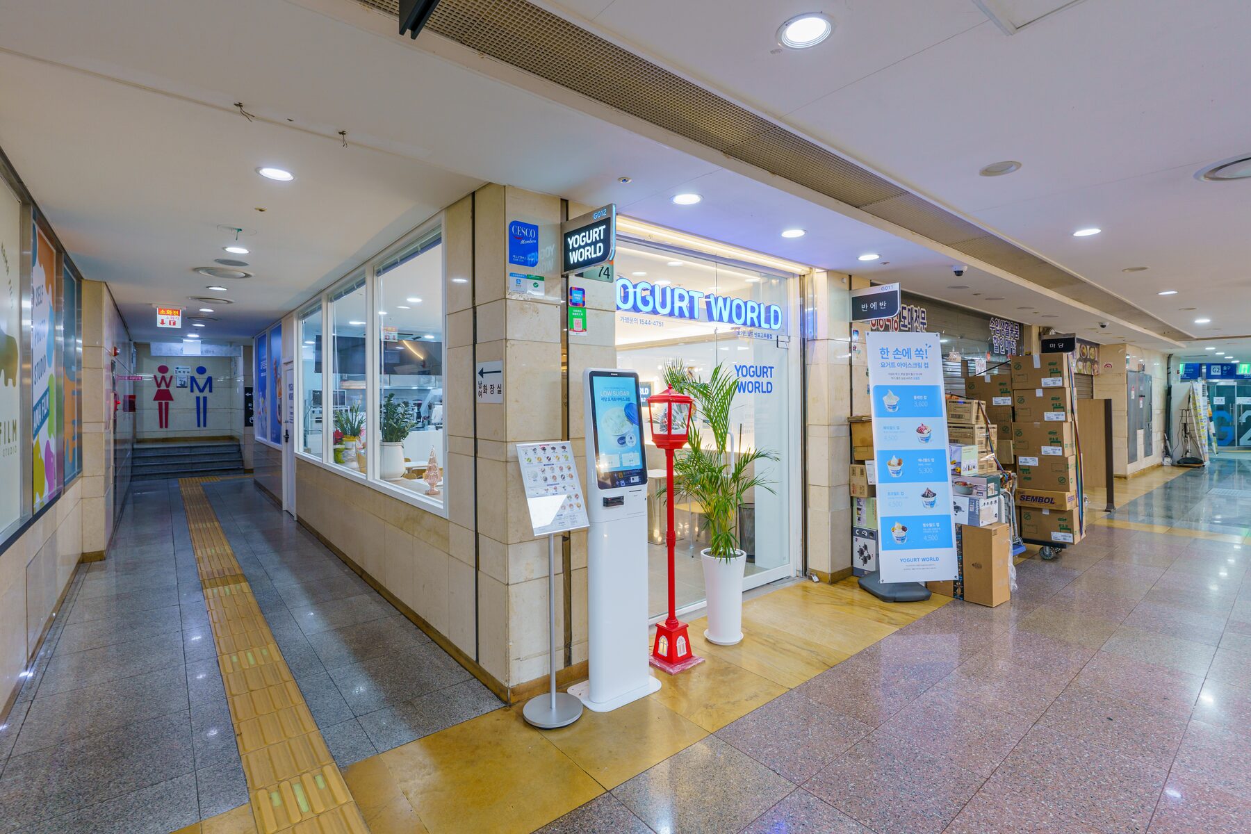 통로 모퉁이 유리 매장, 앞에 흰 단말기와 붉은 기둥, 화분